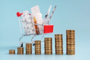 Ascending stacks of coins with a shopping cart filled with medicinal tablets and pills in the background. Represents rising drug prices or costly medications.