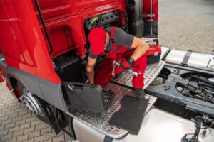 White male heavy-duty vehicle engineer sitting on the back of a red semi-truck, retrieving tools from a toolbox to begin repairs.