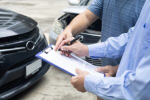 Insurance officer writing on a clipboard while an agent examines a black car after an accident