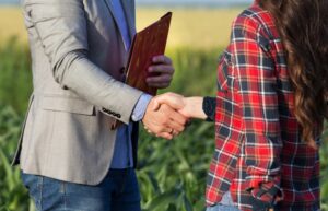 Female farmer shaking hands with an insurance adjuster holding a folder in a cornfield.