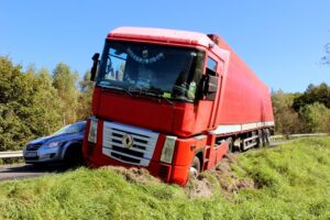 A red semi-truck is stuck in a grassy ditch beside a road, with a silver car positioned behind it.